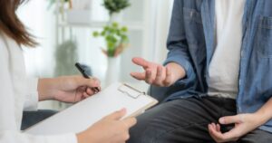 - Treat Mental Health Washington Counselor taking notes on a clipboard while a client gestures during a therapy session.