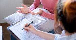 - Treat Mental Health Washington Therapist writes on a clipboard as a client gestures with open hands during a counseling session nearby notebook and pillow visible.