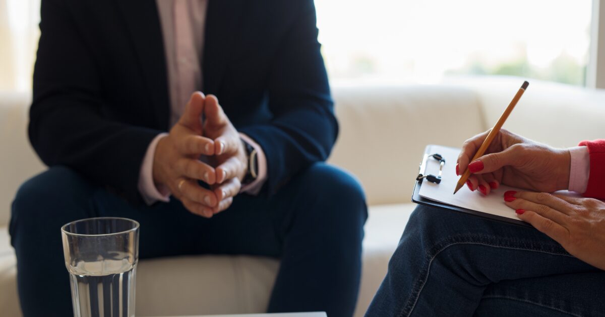- Treat Mental Health Washington Therapy or counseling session: person in a suit sits with hands clasped while another person writes on a clipboard.