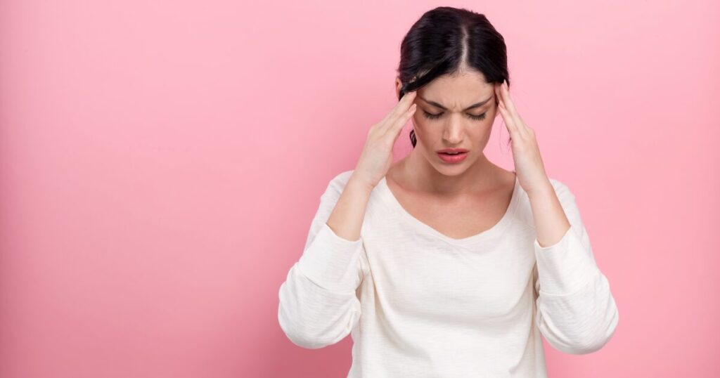 Woman with dark hair in a white top holds her temples and looks stressed against a pink background, suggesting a headache or tension.