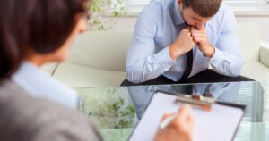 - Treat Mental Health Washington Man in a light blue shirt sits on a sofa, hands clasped under his chin, as a therapist in the foreground takes notes on a clipboard.