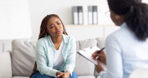 - Treat Mental Health Washington Woman on a sofa during counseling, talking with a therapist who is taking notes on a clipboard.