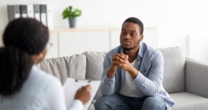 - Treat Mental Health Washington Man in casual shirt sits on a sofa during a therapy session, hands clasped, listening attentively to a clinician taking notes off-camera