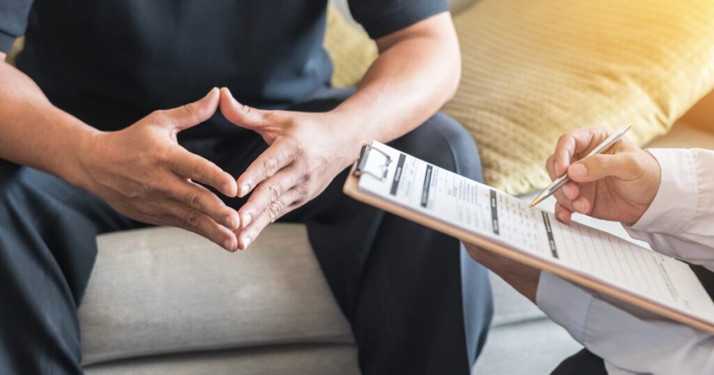 Patient sits on a couch with hands folded together, while a clinician takes notes on a clipboard nearby, indicating a medical or counseling consultation.