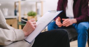 - Treat Mental Health Washington Person taking notes on a clipboard during a therapy or counseling session with a second person seated nearby, visible in the background in a cozy room.