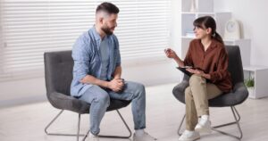 - Treat Mental Health Washington Counselor with a clipboard talks to a man during a counseling session in a bright, minimalist office.